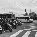 Black and white image of a city intersection with scooters and buses at a crosswalk.