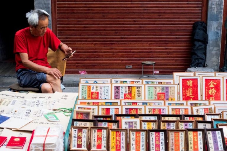 Elderly man in red shirt sitting outdoors, browsing art prints in an urban market.