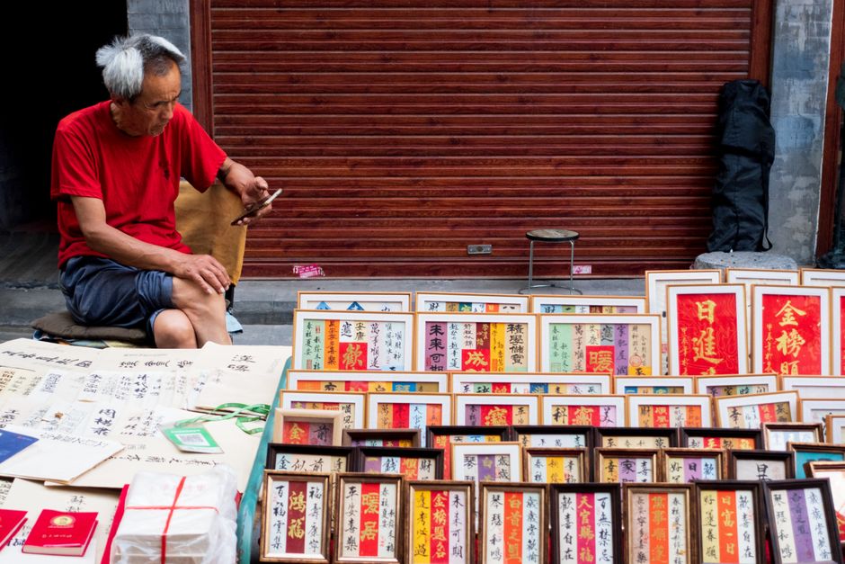 Elderly man in red shirt sitting outdoors, browsing art prints in an urban market.