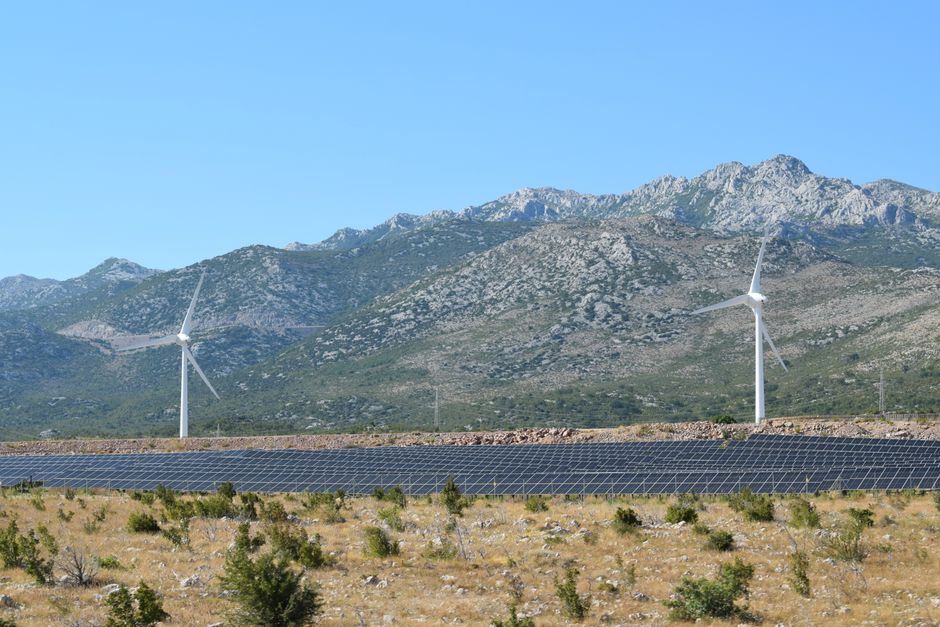 A scenic view of windmills and solar panels against mountainous terrain under a clear blue sky.