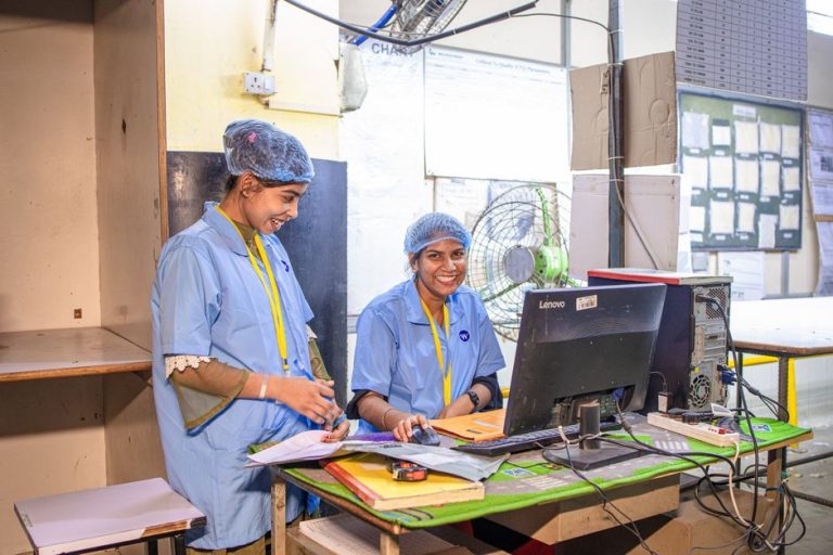 Two female textile workers smiling at their desk in a factory setting.
