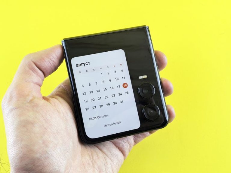 Close-up of a hand holding a flip smartphone with a calendar on screen against a vibrant yellow background.