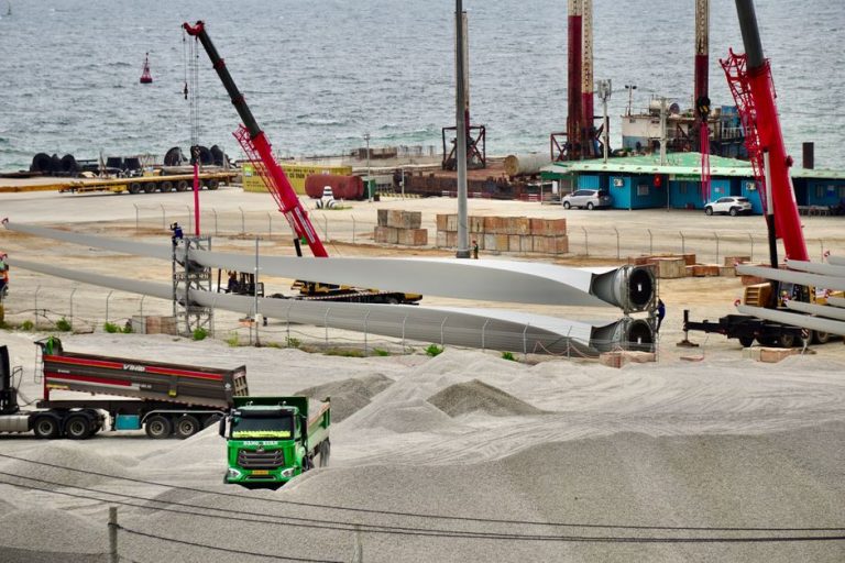 Construction site featuring wind turbine blades, cranes, and trucks by the seaside, showcasing industrial activity.