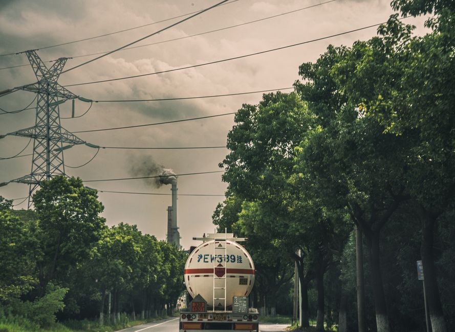 Tanker truck driving on a road by a smokestack emitting smoke under power lines and trees.