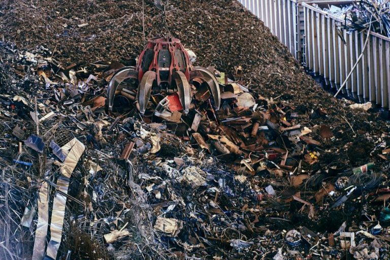 A claw excavator sifting through a large pile of industrial scrap metal outdoors.