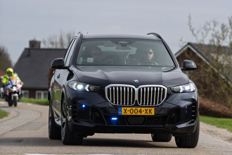 A sleek black luxury SUV travels on a rural road in the Netherlands with a motorbike escort.