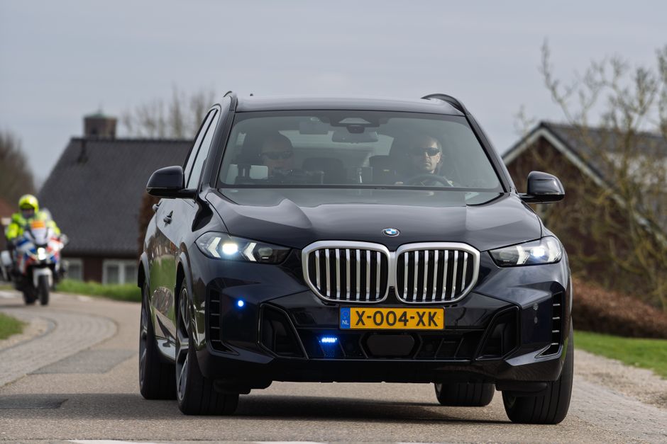 A sleek black luxury SUV travels on a rural road in the Netherlands with a motorbike escort.