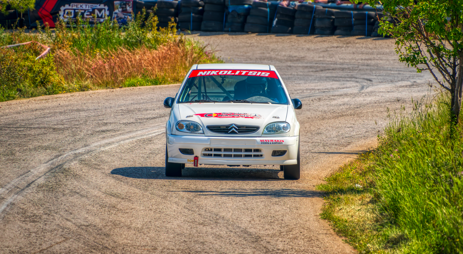 A rally car speeds through a curvy track surrounded by greenery in Ritsona, Greece.