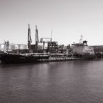 Monochrome photo of a cargo ship docked at an industrial port with cranes in the background.