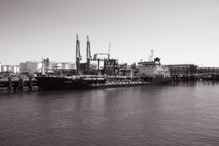 Monochrome photo of a cargo ship docked at an industrial port with cranes in the background.