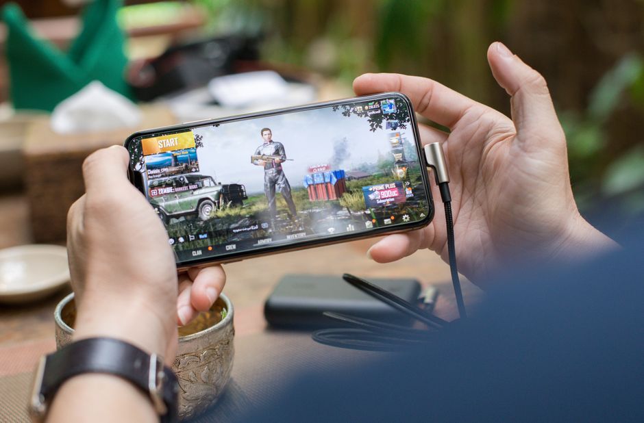 Person playing PUBG on smartphone outdoors with charging cable attached.