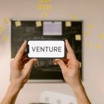 A smartphone displaying 'Venture' held by hands in an office setup. Ideal for business and startup themes.