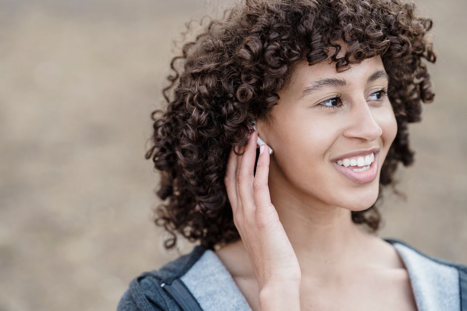 Cheerful ethnic female with black curly hair looking away with smile while listening to songs in wireless earphones on street against blurred background