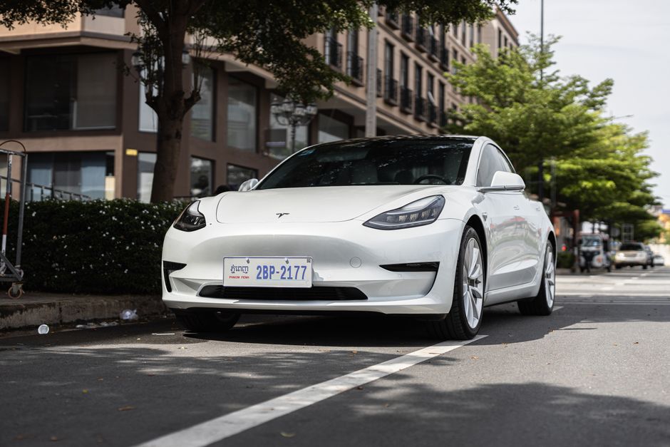 A sleek white Tesla Model 3 parked on a city street under trees, showcasing modern automotive design.