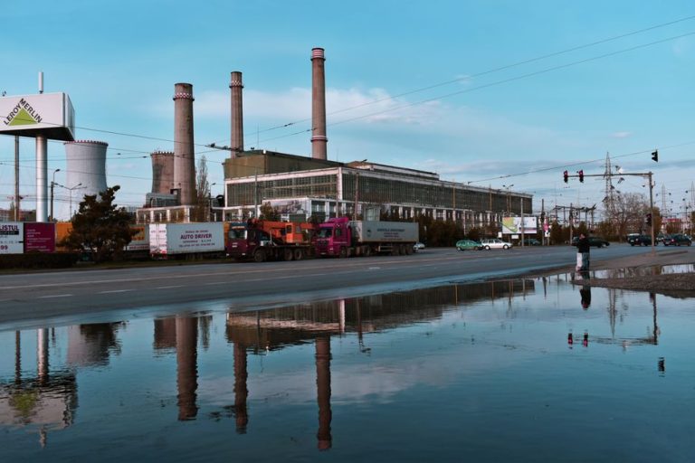 Industrial plant with road and trucks in Iași, Romania, reflecting in water on a sunny day.