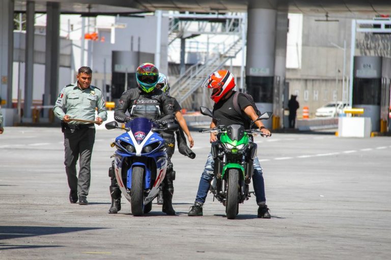 Motorcyclists in helmets at a border checkpoint interacting with a police officer outdoors.