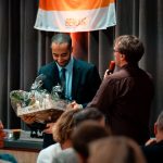 A man receives a gift basket during an award ceremony indoors, creating a celebratory atmosphere.