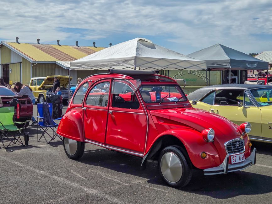 Classic red Citroën 2CV at an outdoor vintage car show with other retro vehicles.