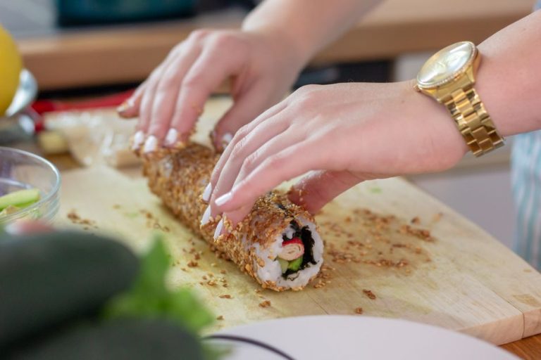 Close-up of hands rolling sushi covered in sesame seeds on a wooden board.