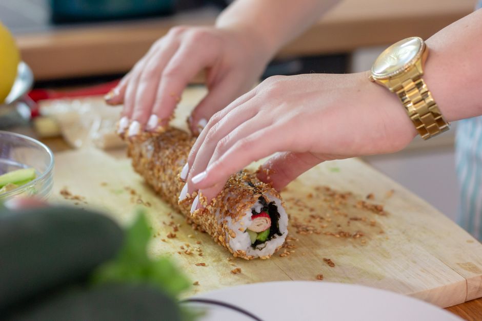 Close-up of hands rolling sushi covered in sesame seeds on a wooden board.