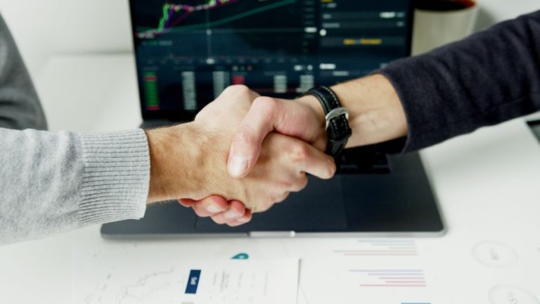 Close-up of a handshake with financial graphs on laptop screen, symbolizing a successful agreement.