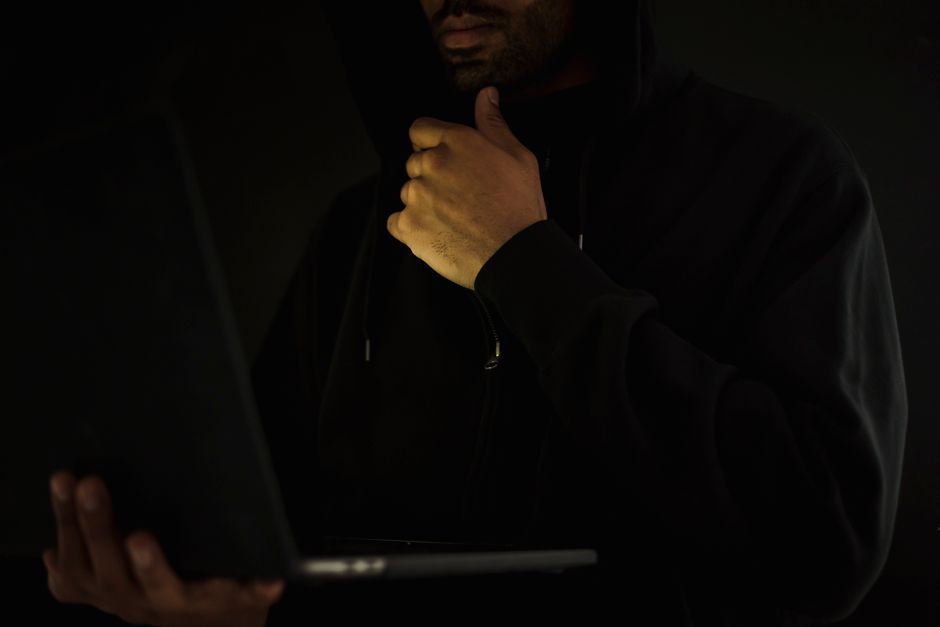 Crop contemplative male hacker in black hoodie watching netbook and touching chin thoughtfully in dark room