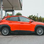 Bright orange SUV parked at a gasoline station with a red truck in the background.