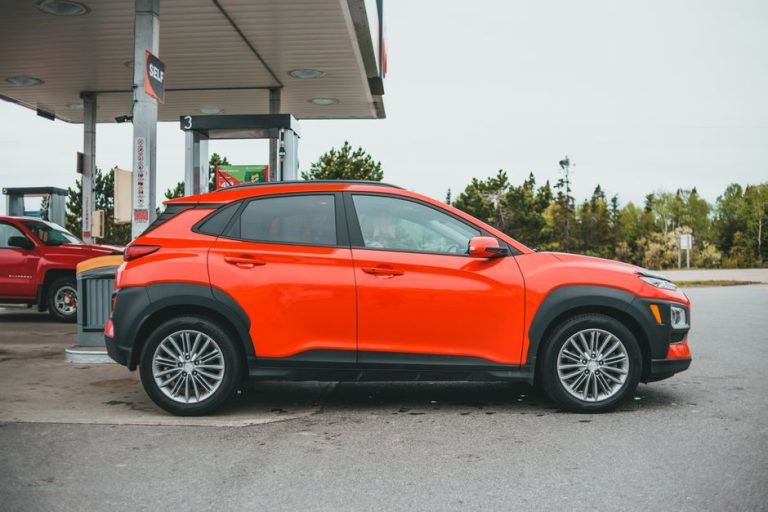 Bright orange SUV parked at a gasoline station with a red truck in the background.