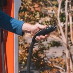 Hand holding an electric vehicle charger at a station outdoors with autumn trees.